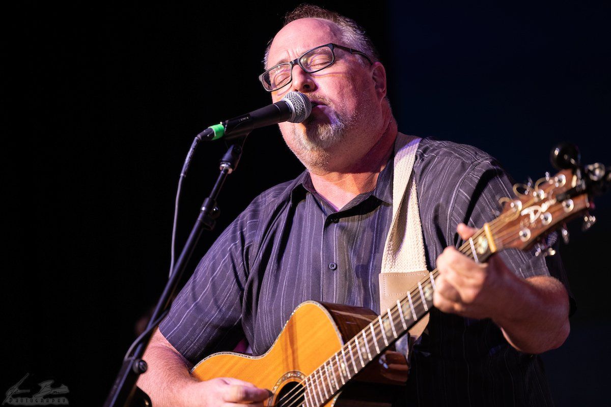 Man playing acoustic guitar, singing into a microphone on stage, wearing glasses and a button-down shirt.