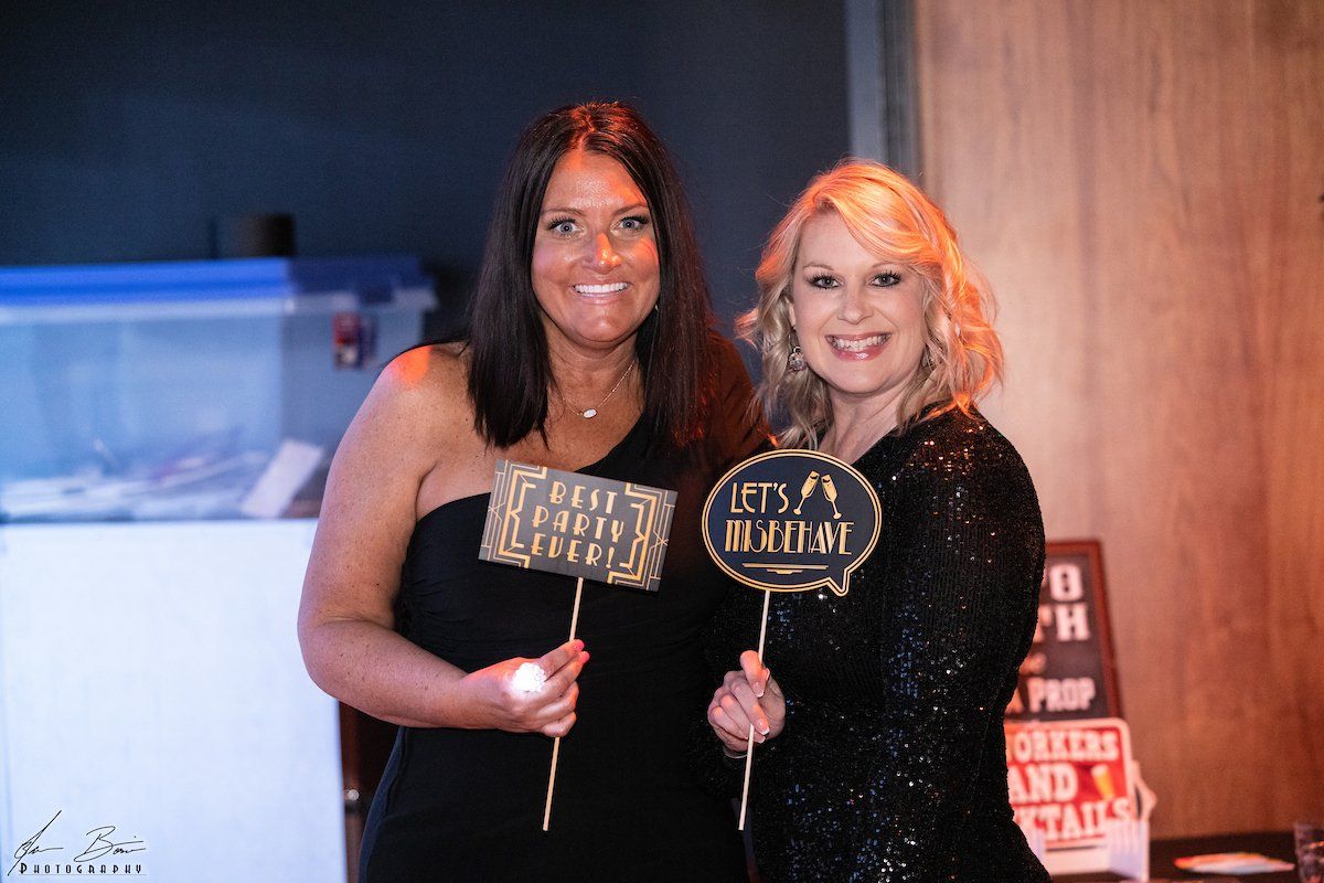 Two women in formal wear smiling, holding photo booth props at an event.
