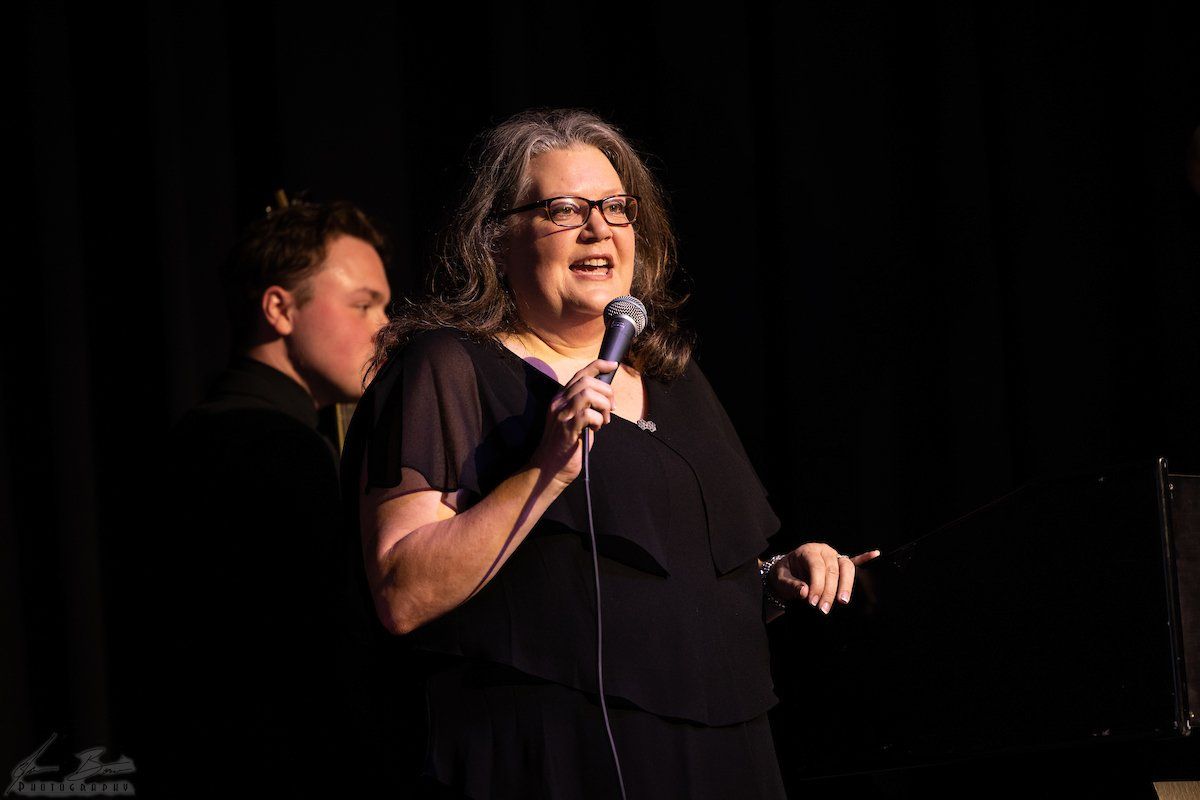 Woman with gray hair, glasses, and a microphone on stage singing, with a dark-suited man to her left.
