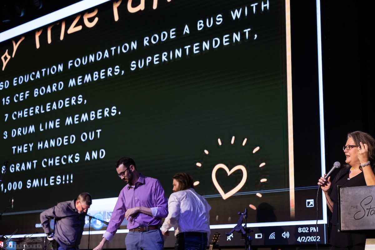 People on stage with a presentation showing a bus ride with a foundation, cheerleaders, and smiles.