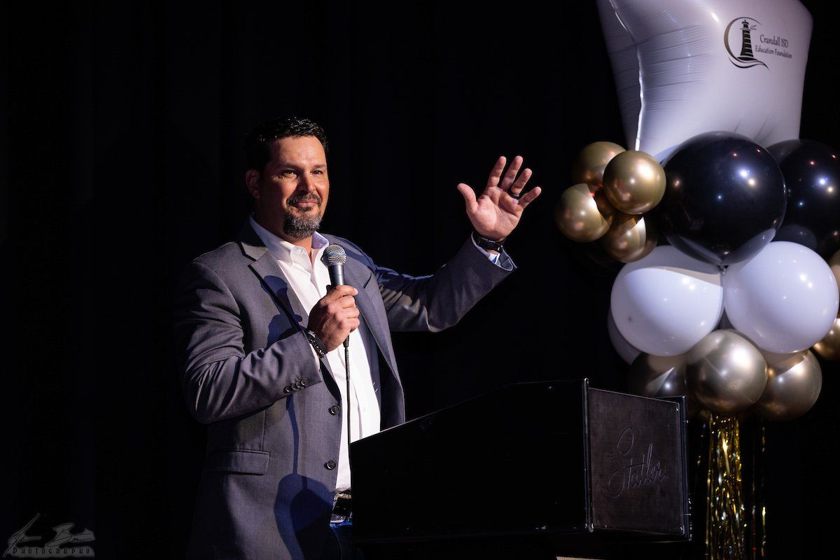 Man in suit speaks at podium, gesturing, with balloons.