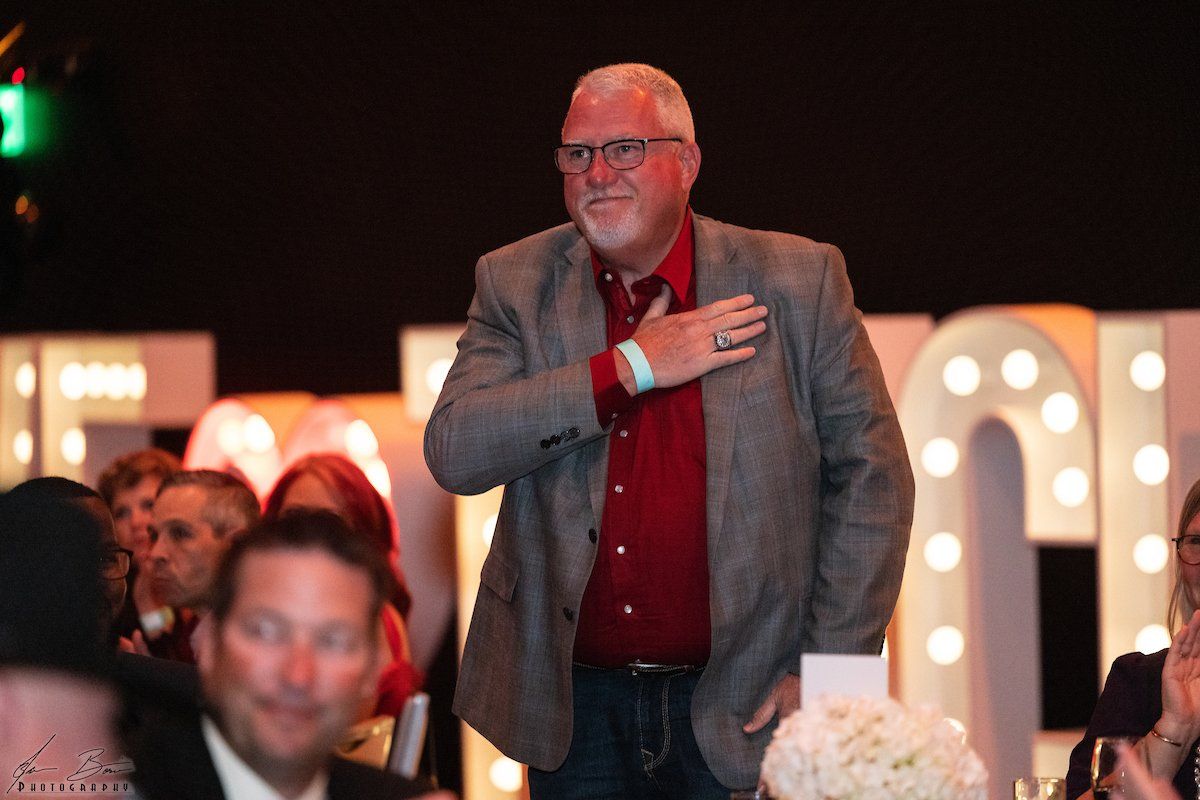 Man in a gray blazer and red shirt, hand on chest, smiling. Event backdrop with large marquee letters.
