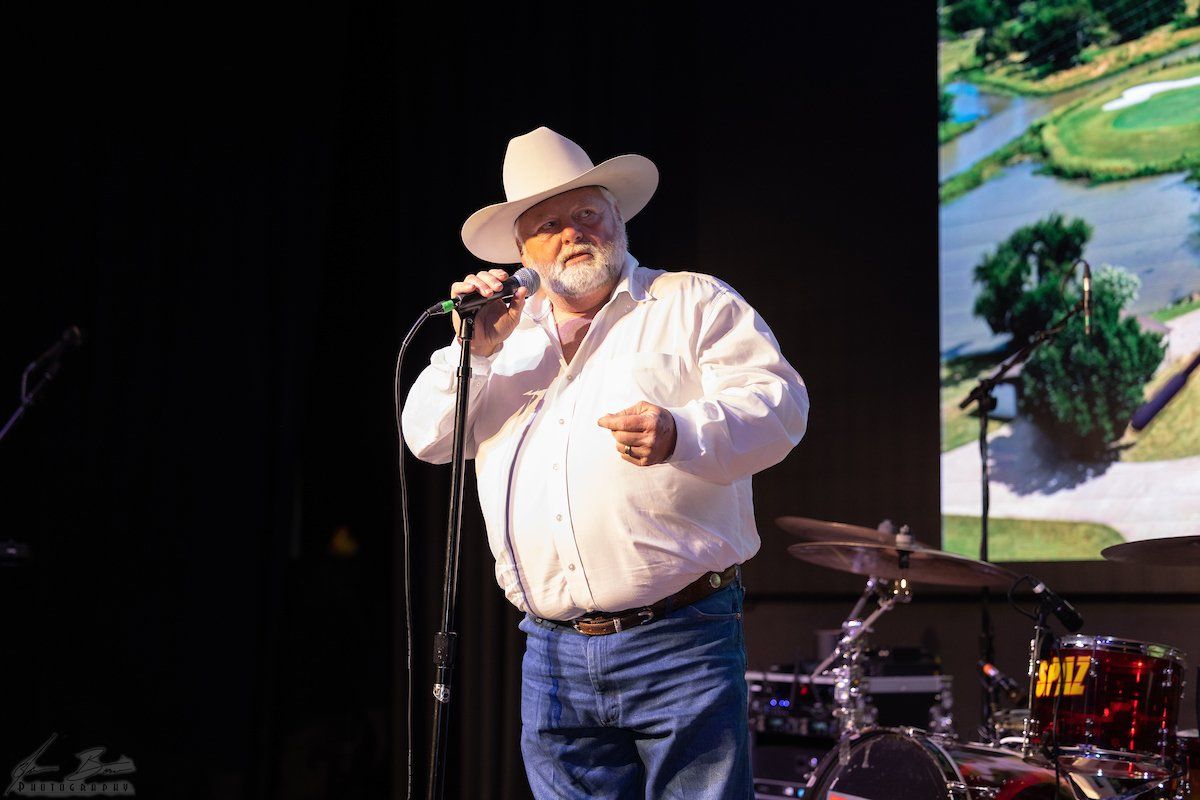 Man in cowboy hat singing into a microphone on stage; behind him a screen with a golf course image.