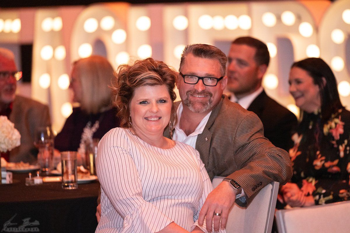 Couple smiling together at a lit event, man in jacket, woman in striped shirt, others at a table.