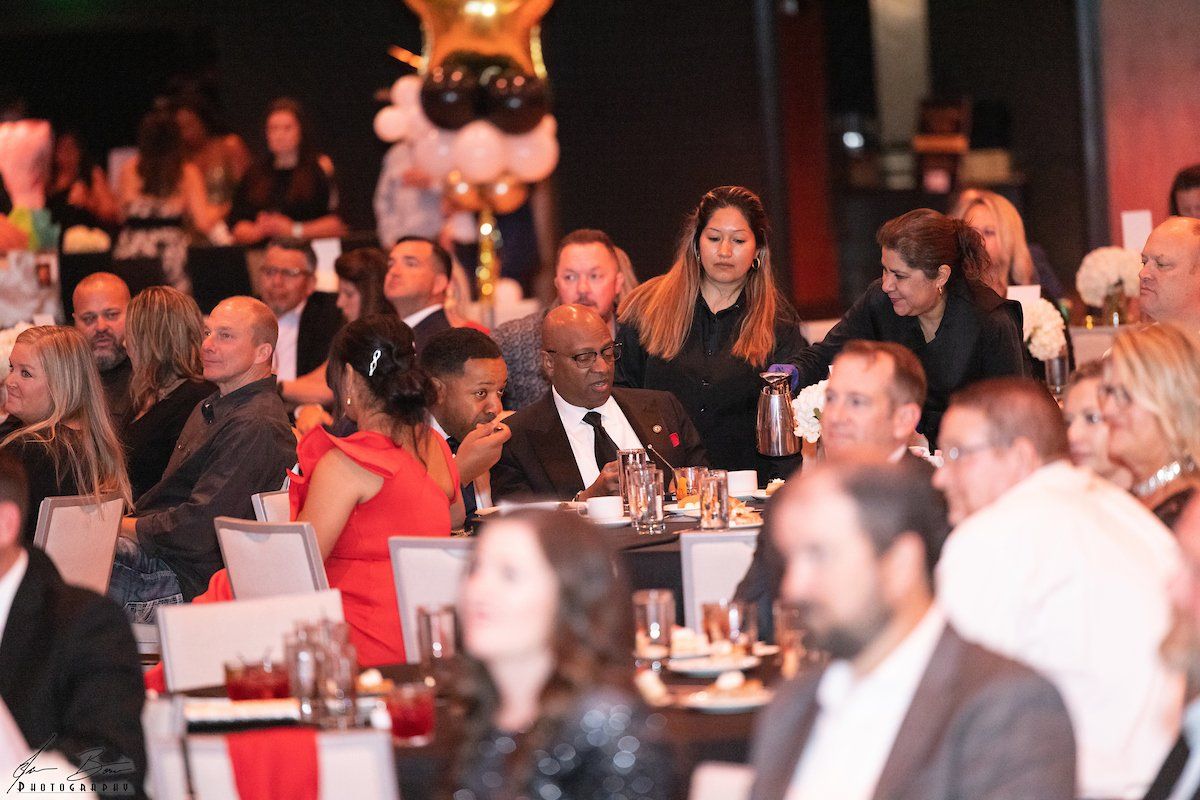 People seated at tables at an event, being served drinks by staff, with red and black decor.