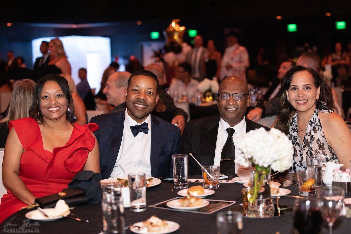 Four people smiling at a table at a gala. Woman in red dress, men in suits, and woman in a patterned top.