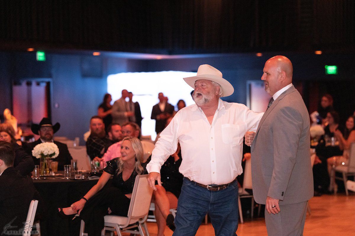 Man in cowboy hat talks with another man in a suit at an event, people and tables in the background.