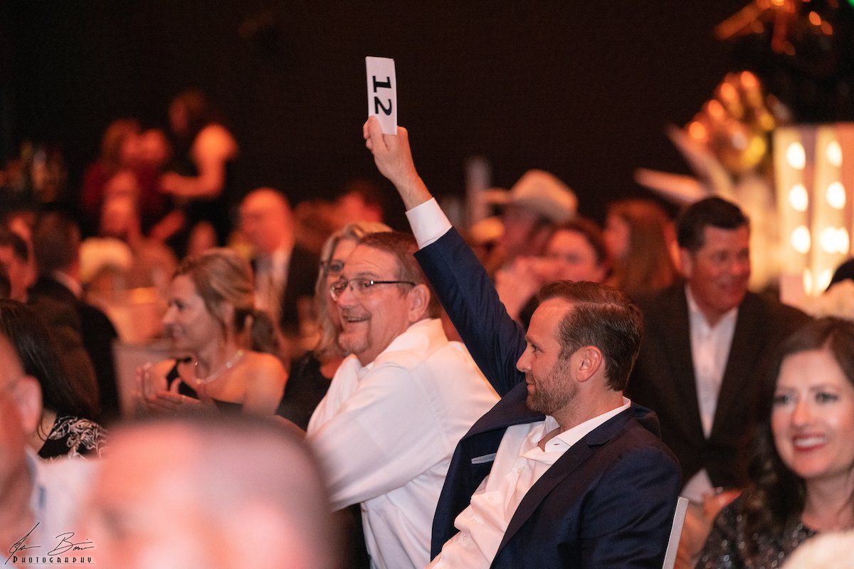 People at an auction, man in blue suit holding up bid card, others in the background watching.