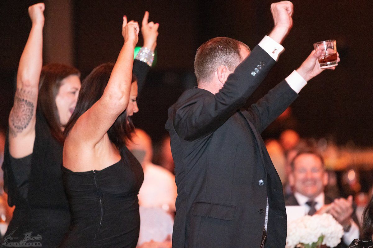 People celebrating at a formal event, raising arms. One man holds a drink, smiling.