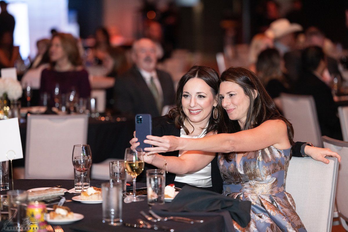 Two women at a formal event take a selfie, smiling. They sit at a table set with wine glasses.