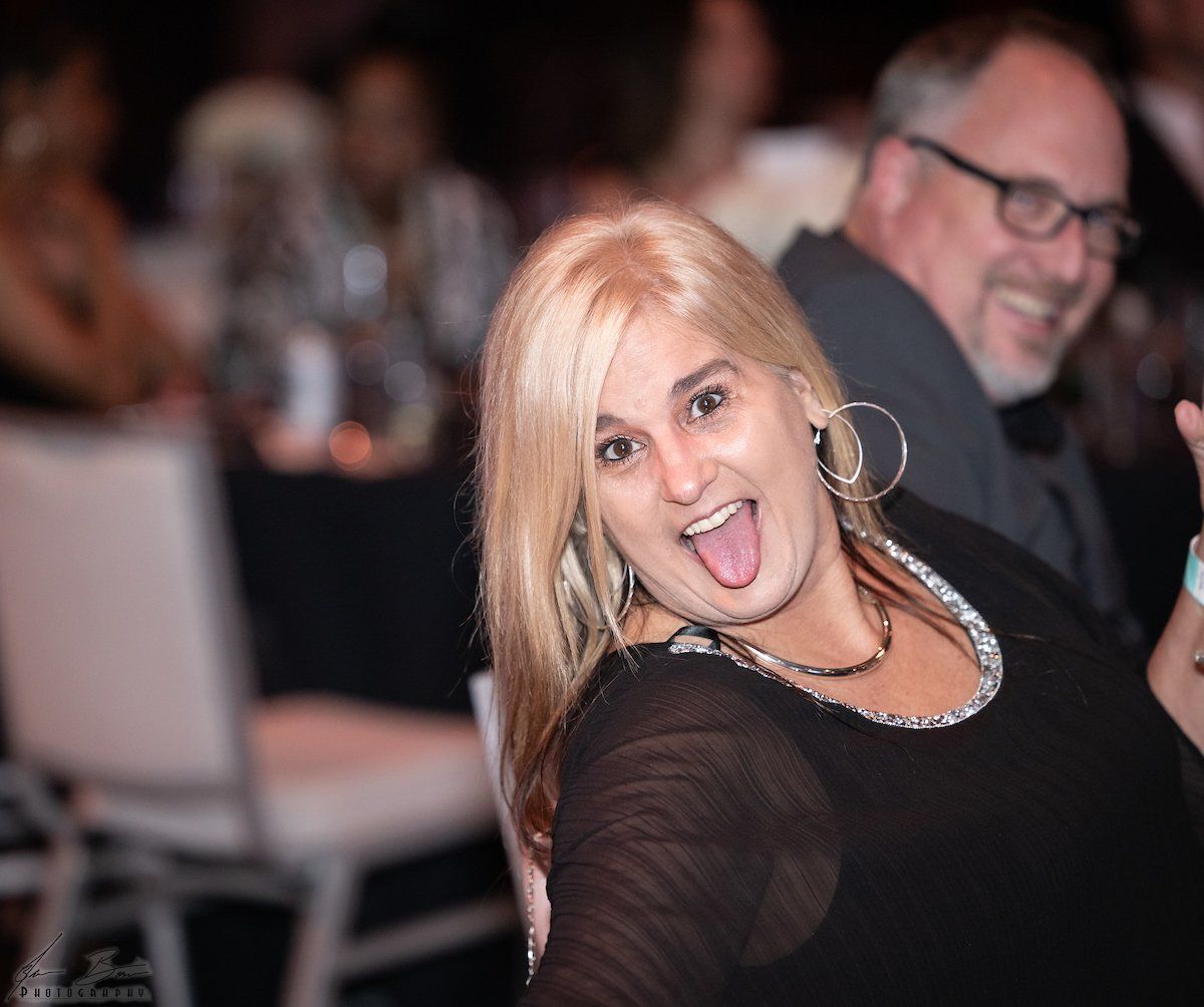 Woman with blonde hair sticking her tongue out at a formal event. Man in background smiles.