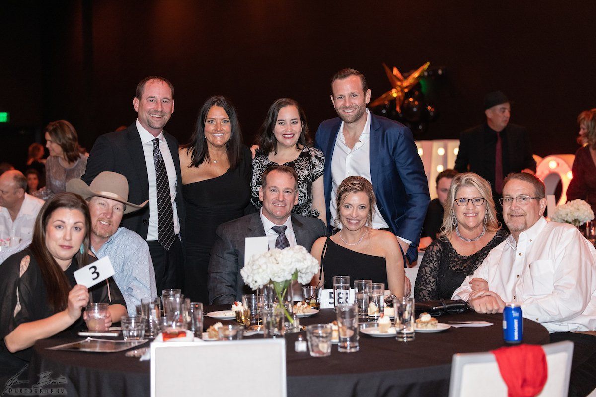 Group photo at a formal event, people smiling, seated at tables. Black and white attire, warm lighting.