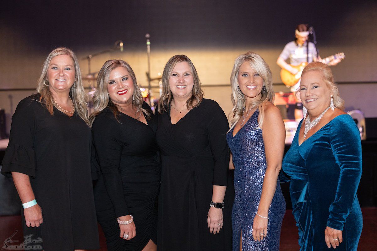 Five women pose, smiling, at an event. The woman in blue velvet dress stands out. A band plays in the background.