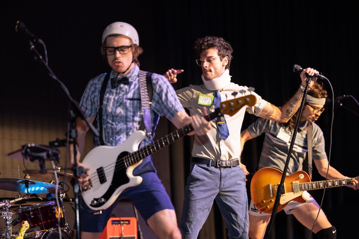 A goofy band performs on stage. One plays bass, another guitar, and a third sings; all wearing silly outfits.
