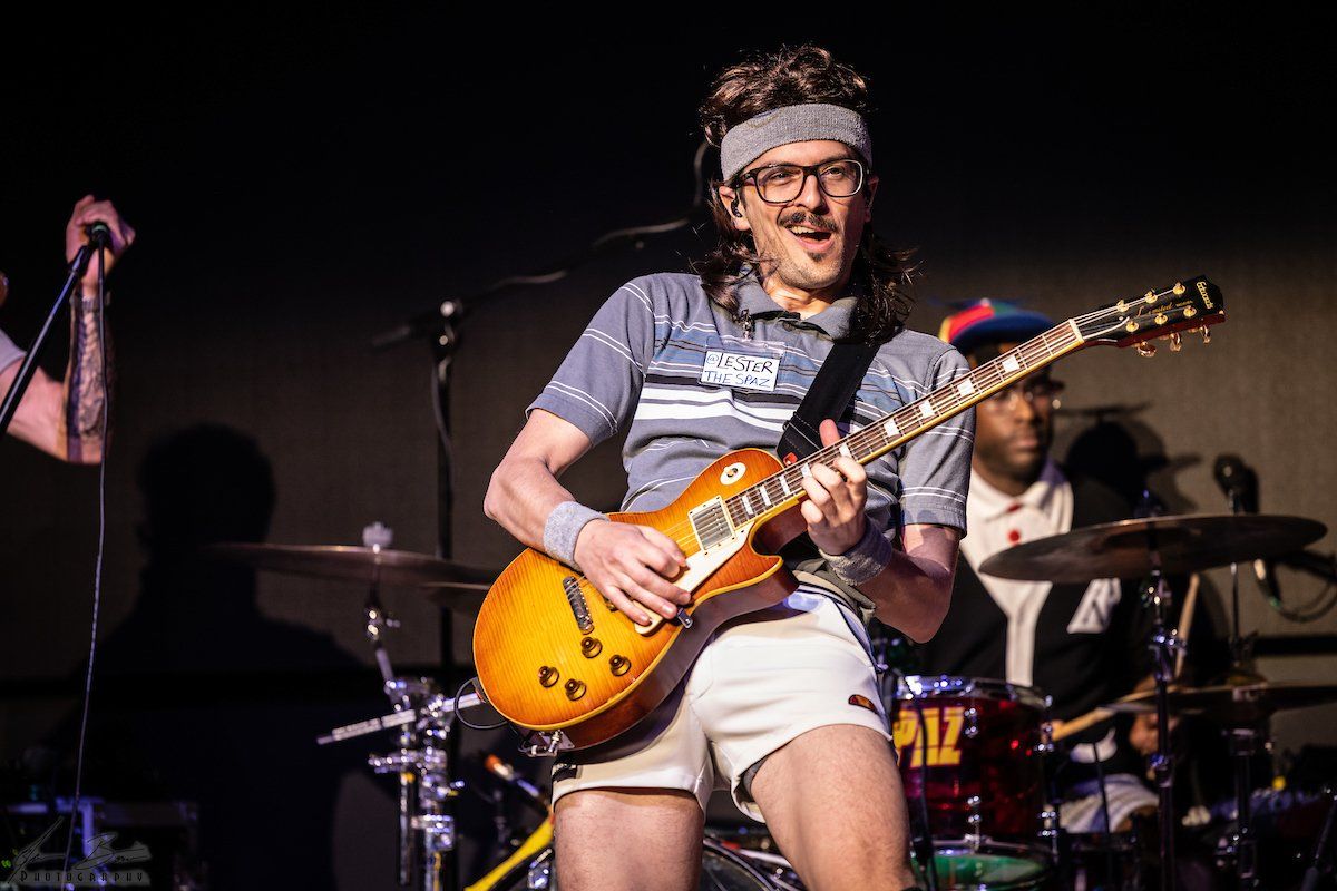 Man playing electric guitar on stage, wearing athletic shorts, headband, and eyeglasses.