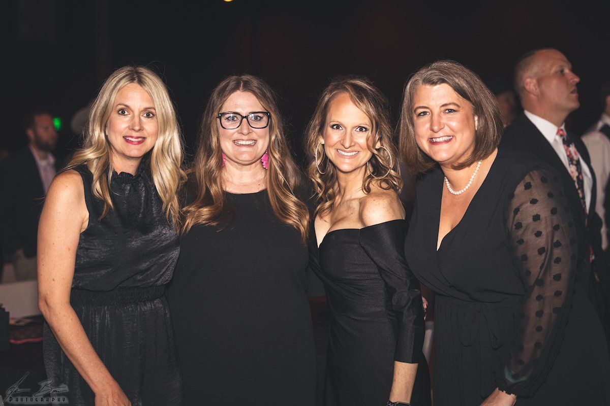 Four women in black dresses pose together at an event.