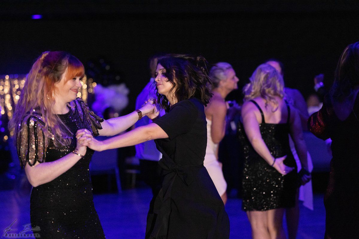 Two women dancing at a party. They are holding hands and laughing, with a black and blue background.