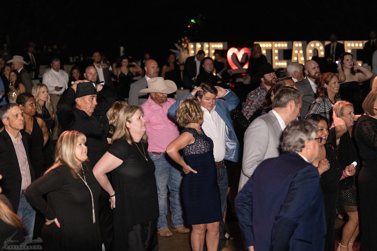 Large crowd at an event; some people talking, others posing. Dark setting with lighted sign.