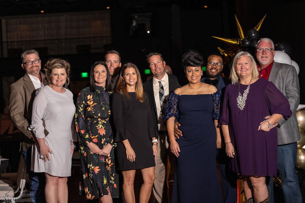 Group of people smiling at an event. Men wear suits and coats; women, dresses. Indoors, dark setting with balloons.