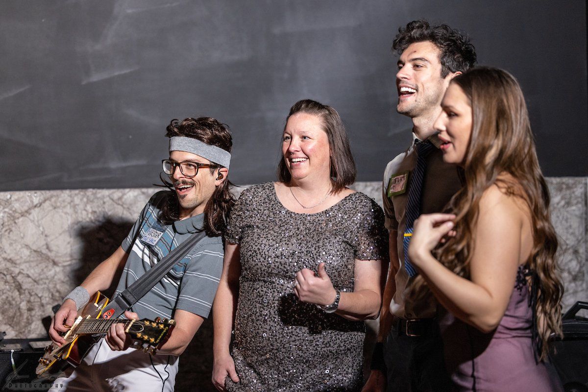A band performs. A man with a headband plays guitar, smiling. A woman and two others sing, smiling and looking up.
