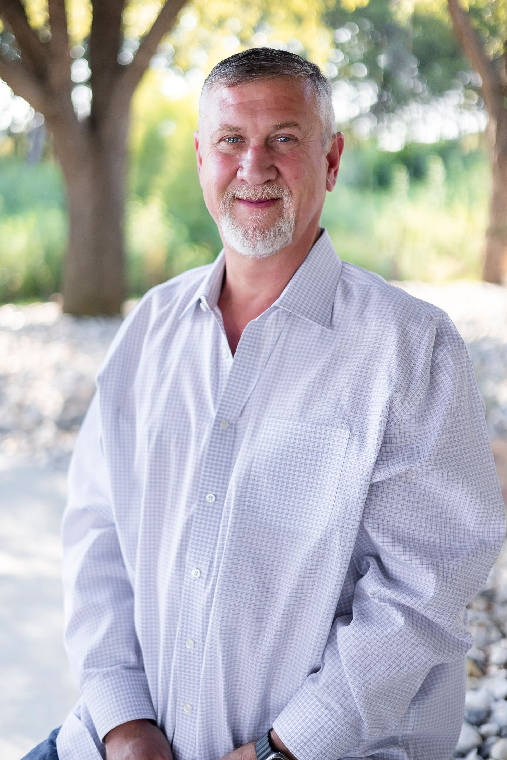 Man with grey hair and beard in a button-down shirt smiles outdoors in front of trees.
