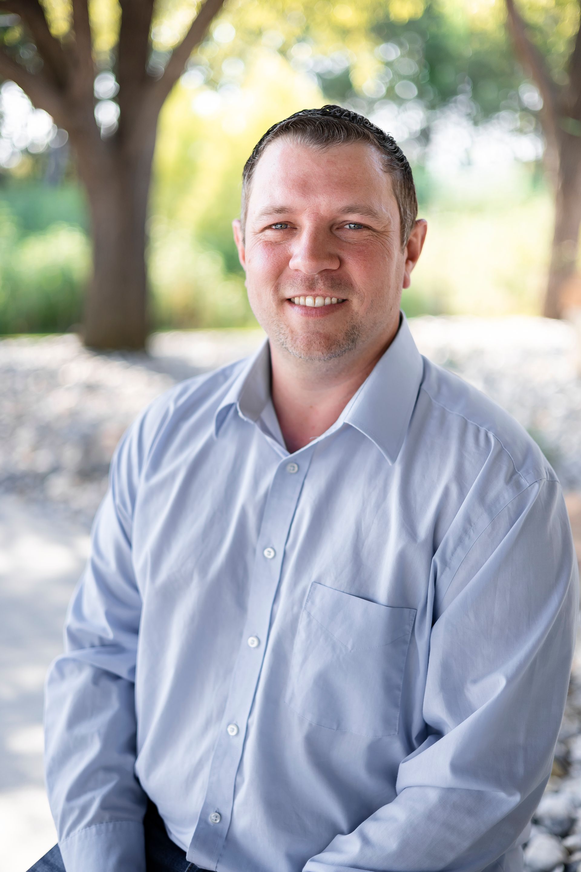 Man in blue shirt smiles outdoors, trees and rocks in background.
