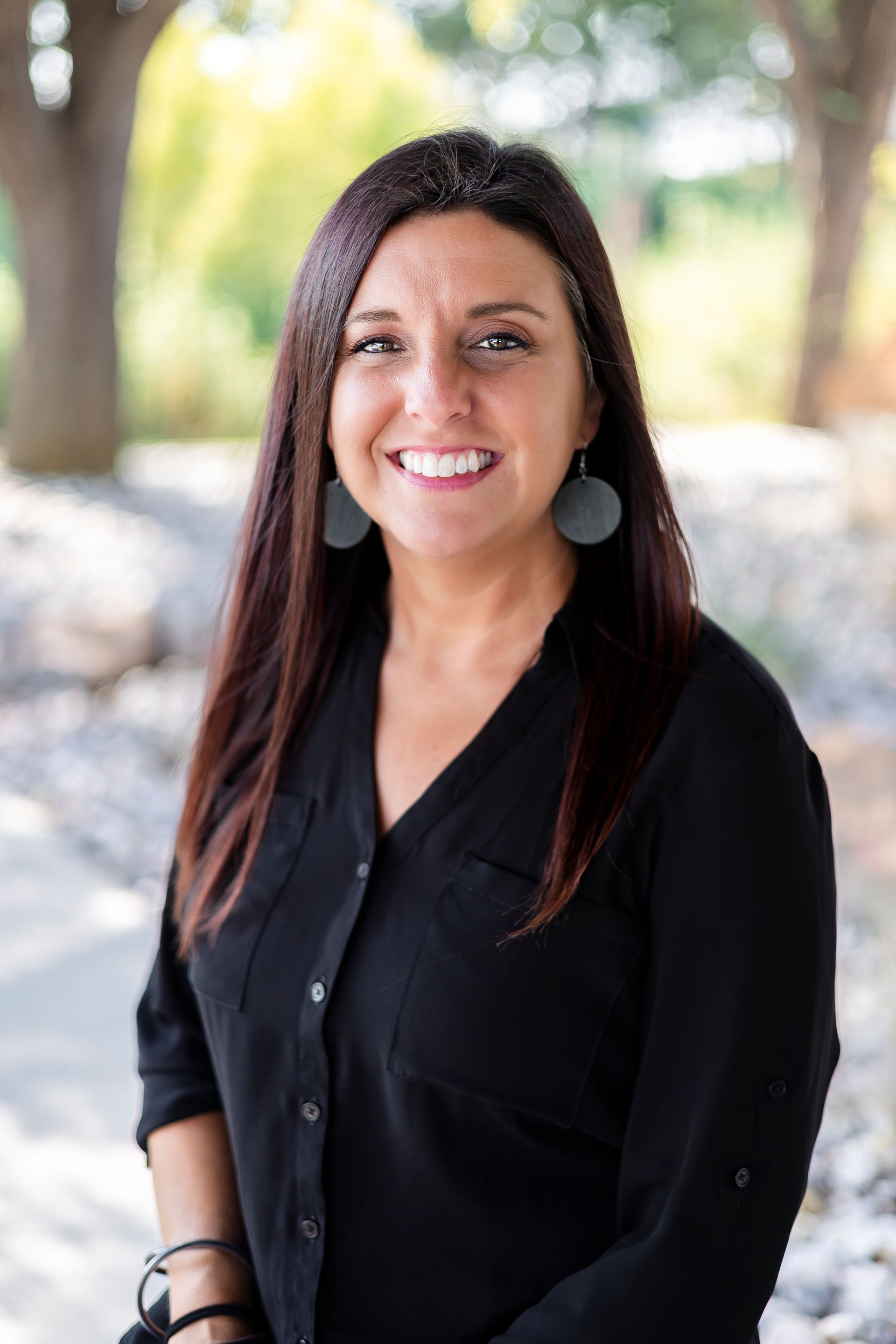 Woman with long brown hair, wearing black shirt, smiling outdoors.