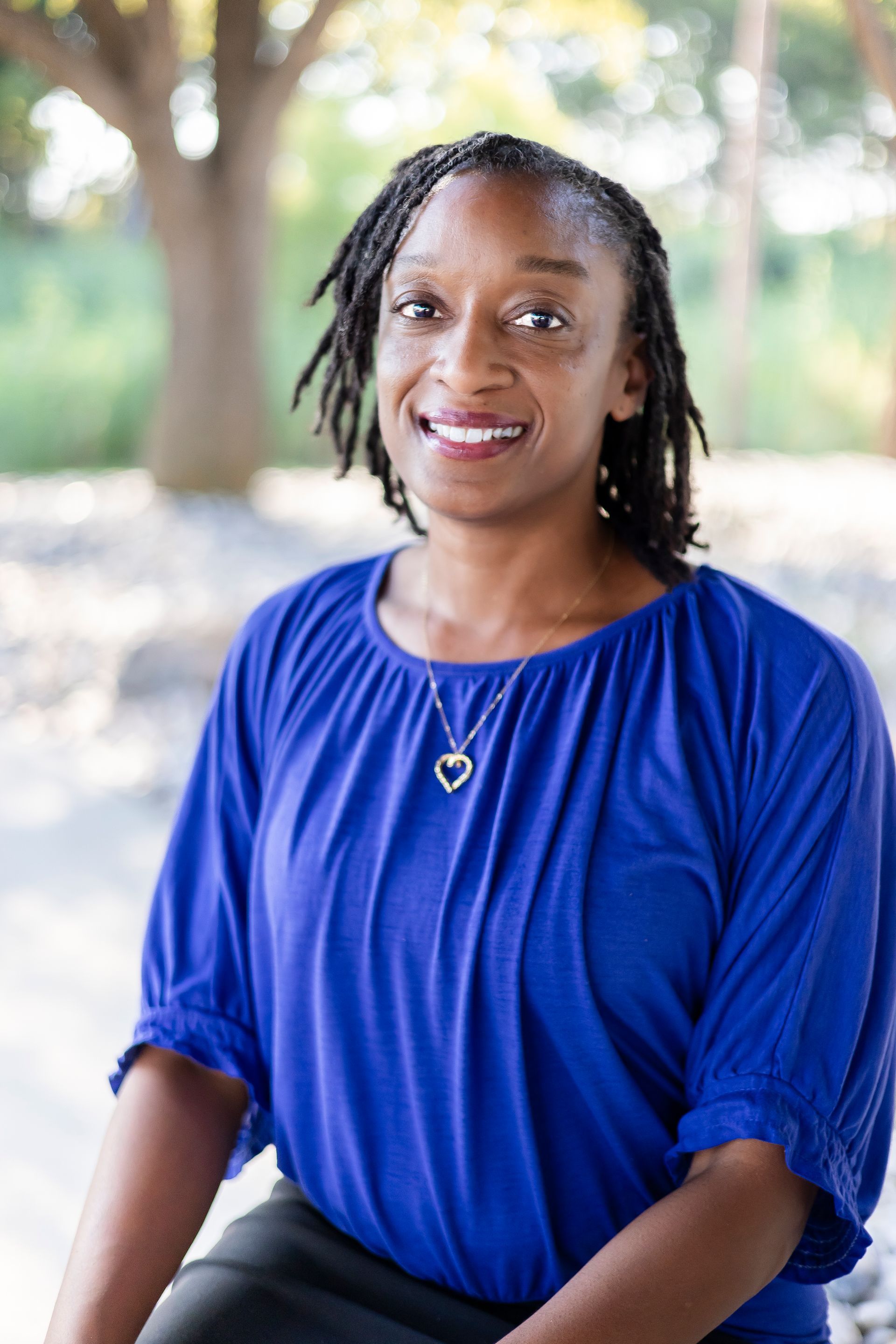 Woman with dreadlocks in blue top, smiling, outdoors with blurred background.