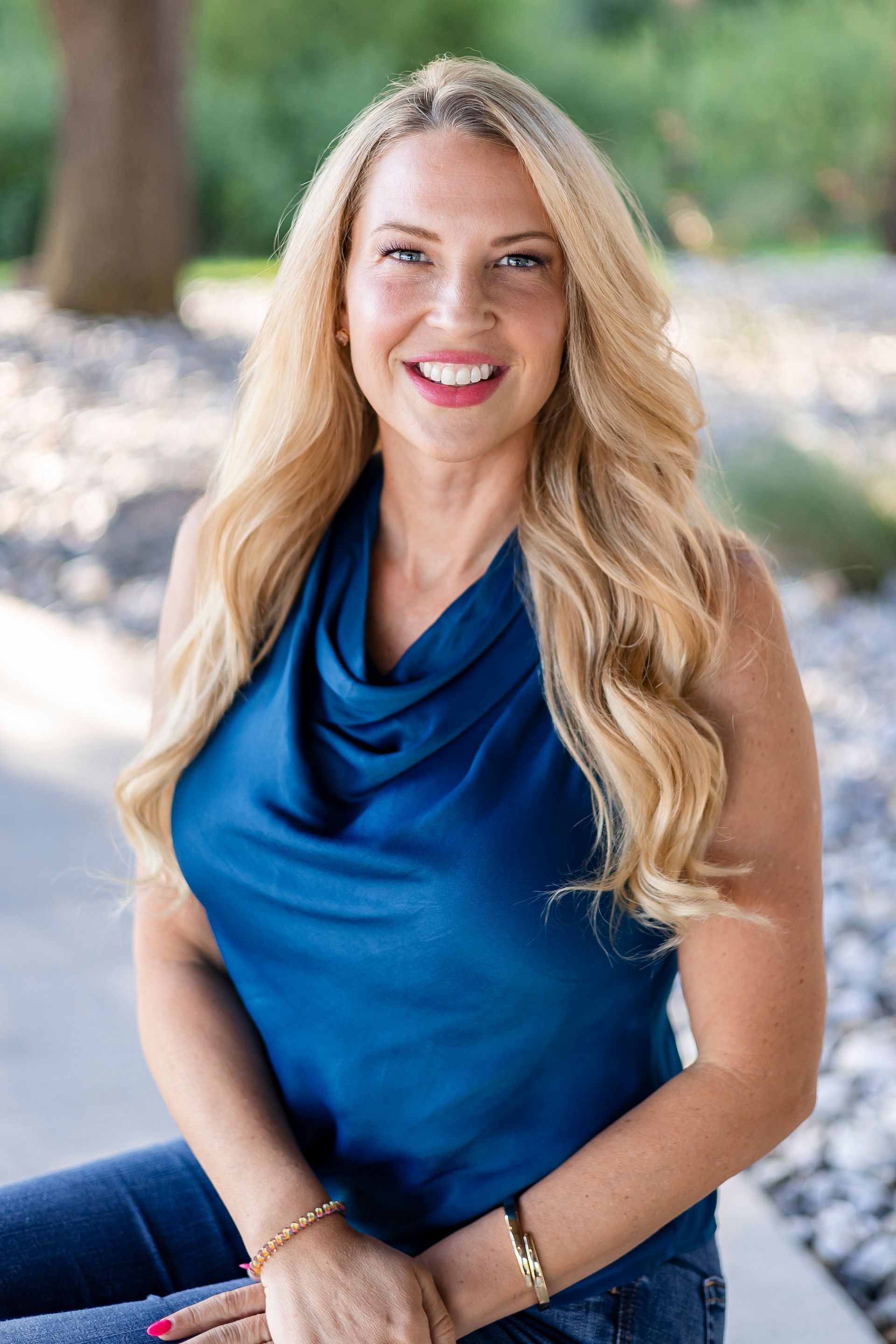 Woman with blonde hair smiles in a blue top and jeans, outdoors.