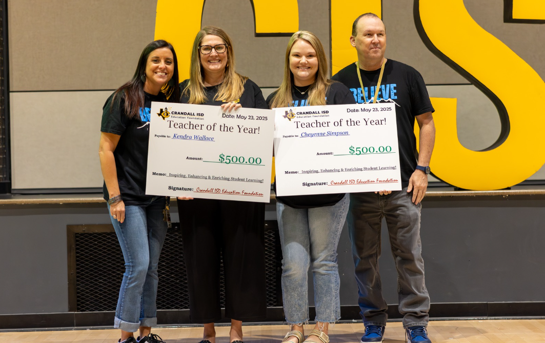 Four people holding oversized checks; in front of a school sign, smiling.