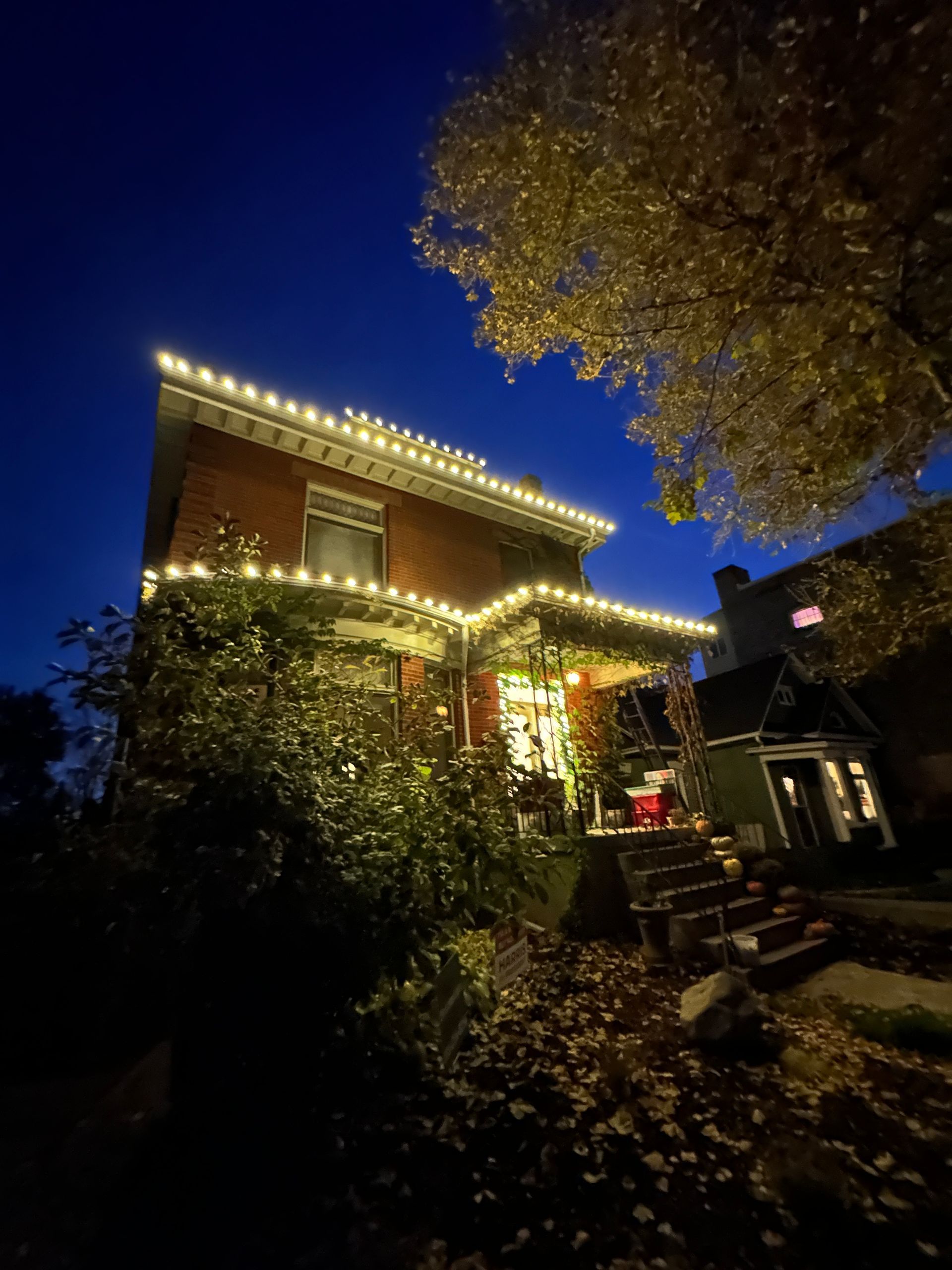 A brick house at dusk with string lights along the roofline and porch, surrounded by trees.