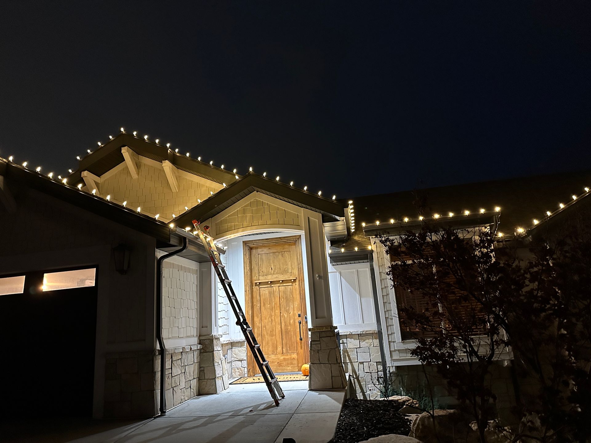 House at night with Christmas lights; ladder propped against the doorway. Garage and snowy ground visible.
