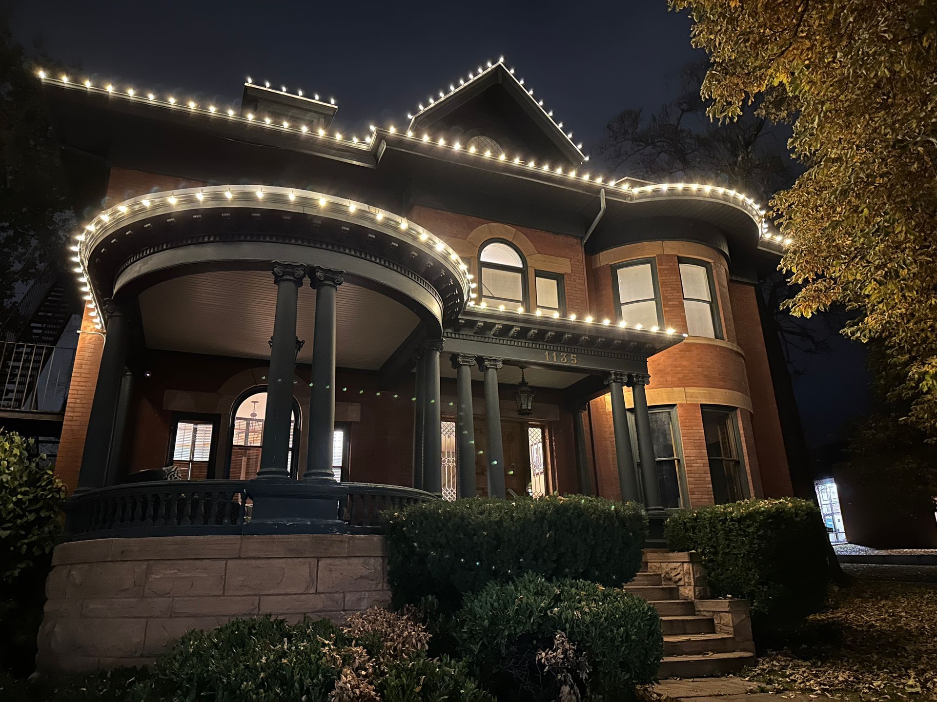 Lit up brick house at night with decorative lights along rooflines and porch.