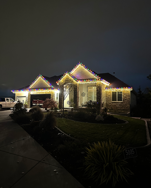 House at night with multi-colored Christmas lights outlining roof, garage, and windows.