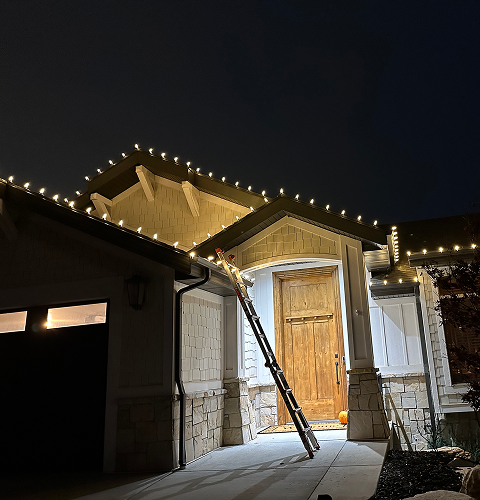 House decorated with lights; a ladder leans against the front door. Nighttime.