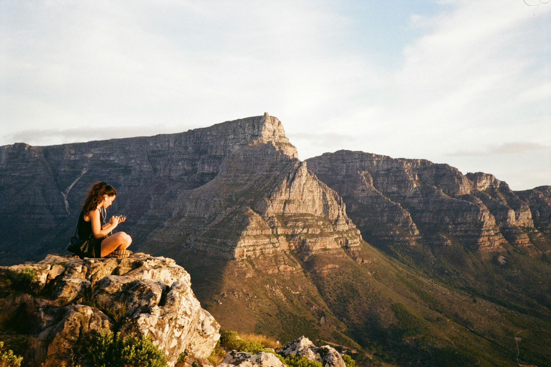 Woman sitting on a rock, looking at mountain range, possibly Table Mountain, in South Africa.