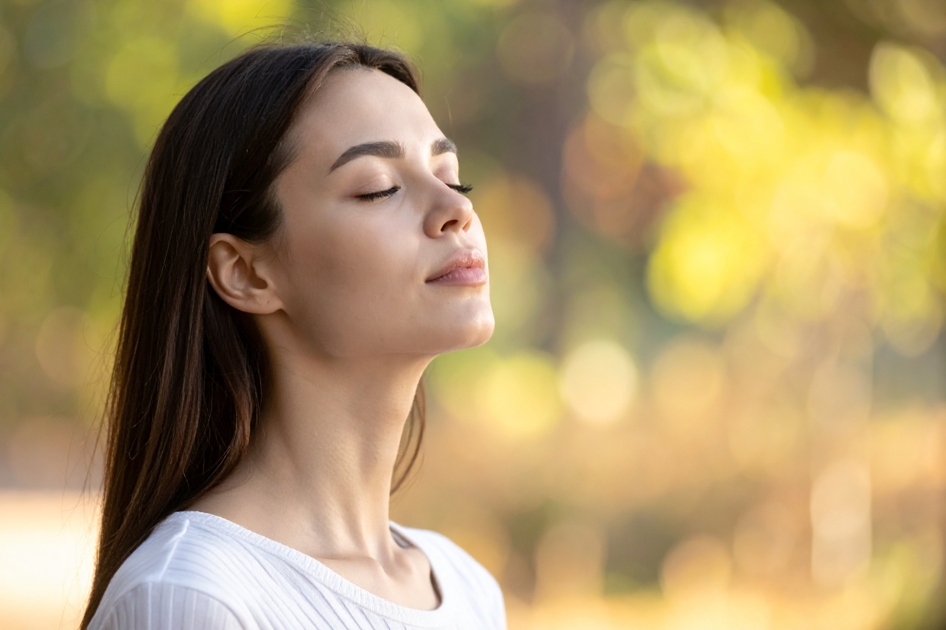 Woman with eyes closed, taking a deep breath outdoors in a sunlit, blurred background.