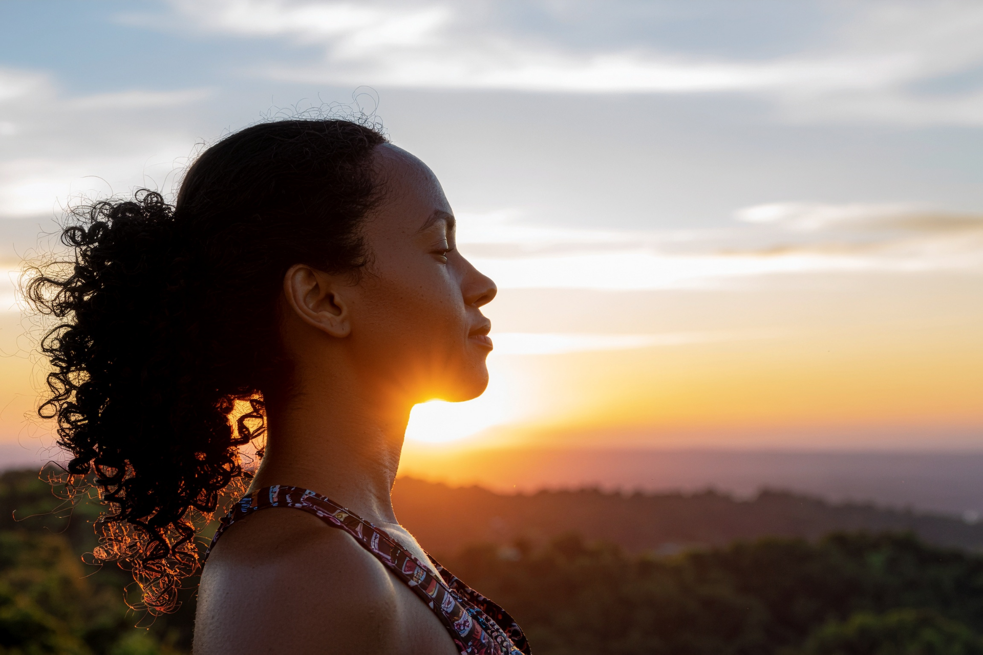 Woman with curly hair looking at sunset, silhouetted against a colorful sky.