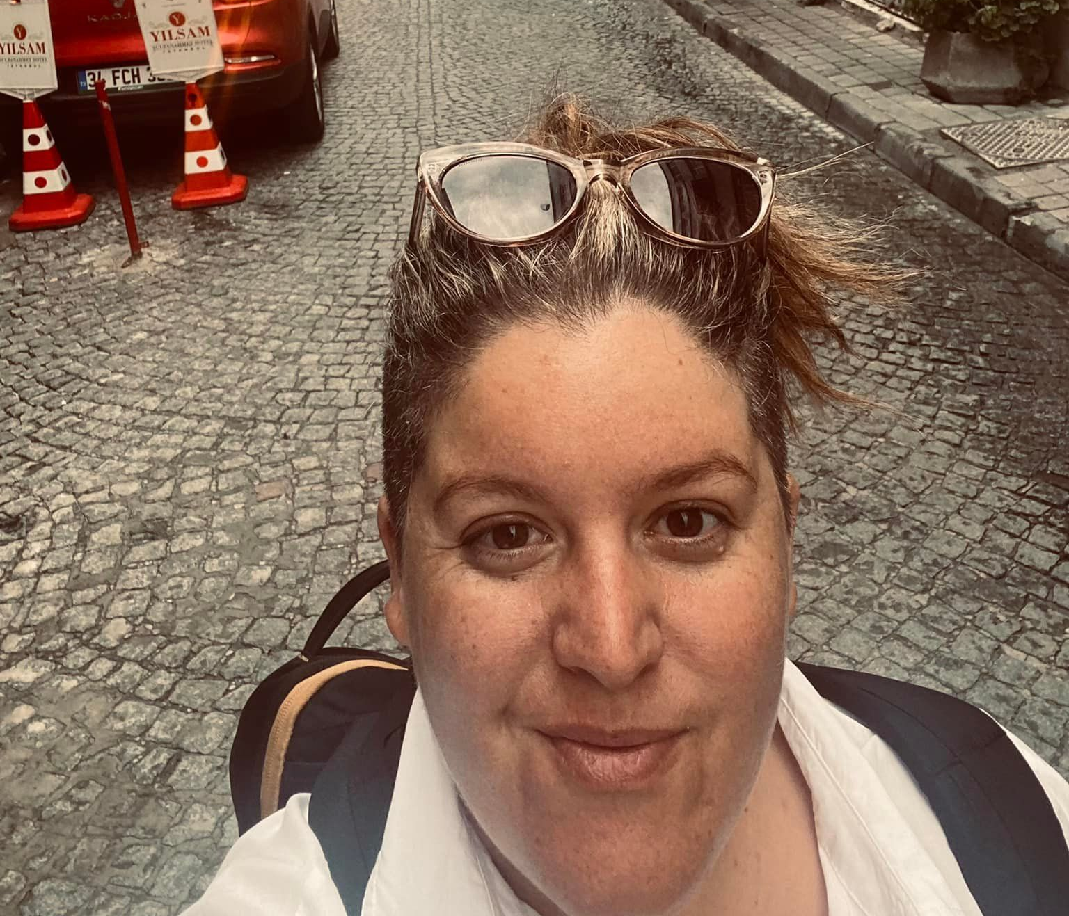 Woman with sunglasses on head takes a selfie on a cobblestone street, backpack visible. Orange car and cones in the background.