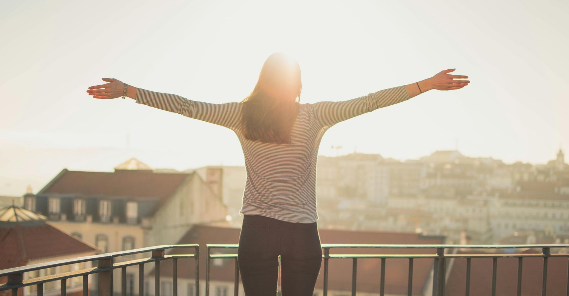 Woman with arms outstretched, standing on a rooftop, silhouetted by the sun, overlooking a city.