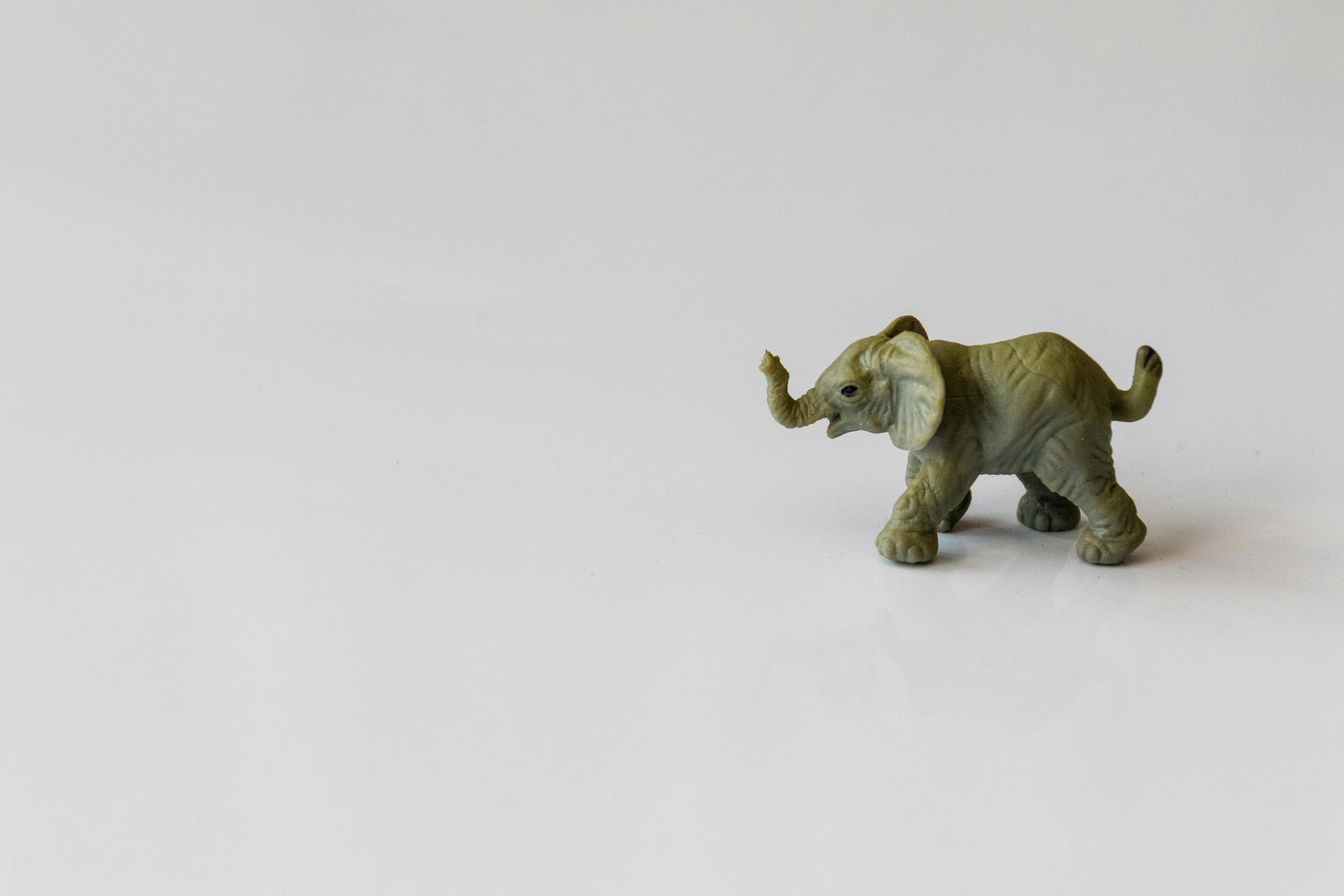 Small green toy elephant on a white surface, trunk raised, walking right.