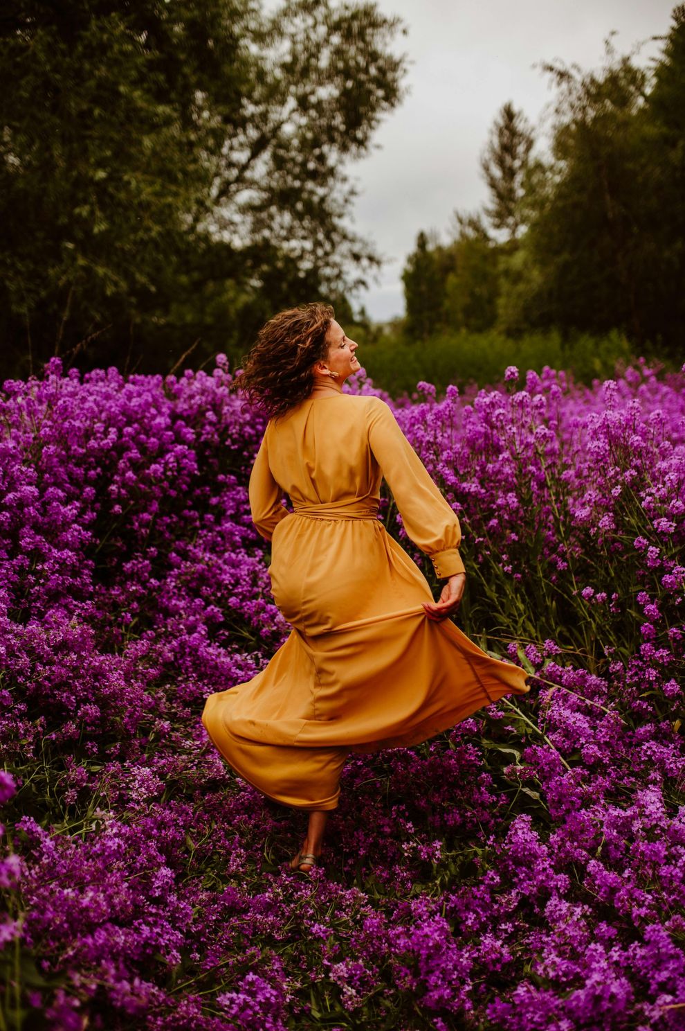 Woman in mustard dress twirling in a field of vibrant purple flowers, looking upward, outdoors.