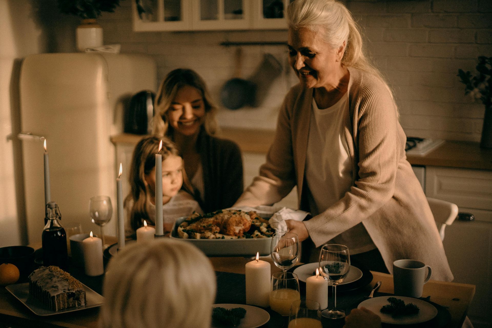 Family gathers around a table with a roast chicken, candles, and a cake in a warmly lit kitchen.