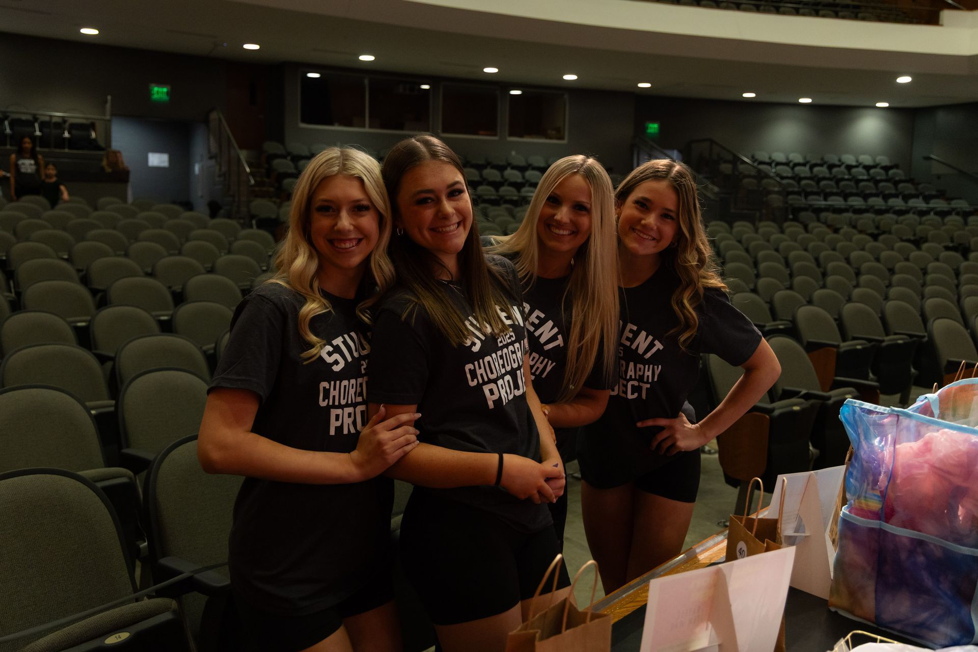 Four young women in matching shirts pose in an auditorium. They smile, and some have hands on each other.