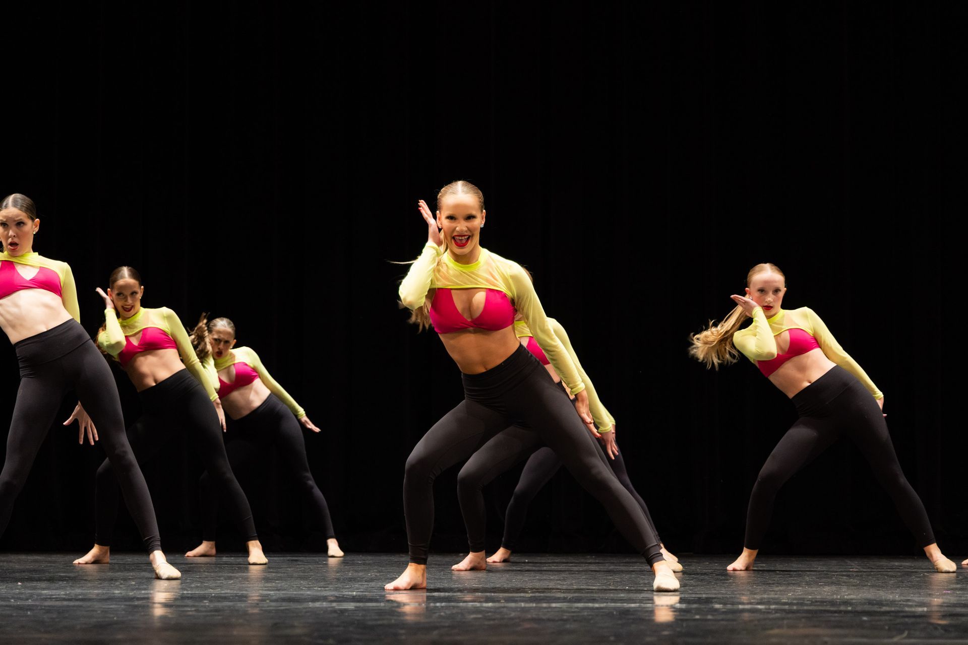 Dancers in pink tops, black leggings, performing on stage. Black background.