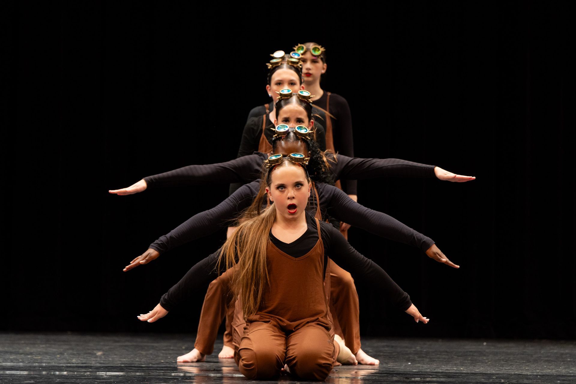 Dancers in formation, black tops and brown overalls, arms outstretched. One dancer has an open mouth. Black background.