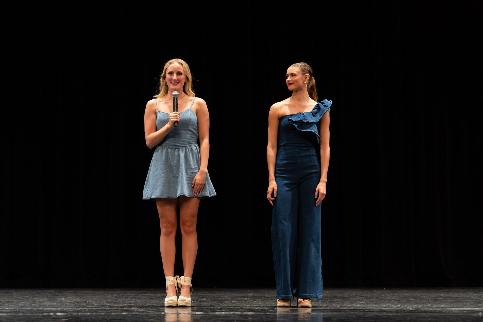 Two women on stage, one holding a microphone and smiling. Dark backdrop.