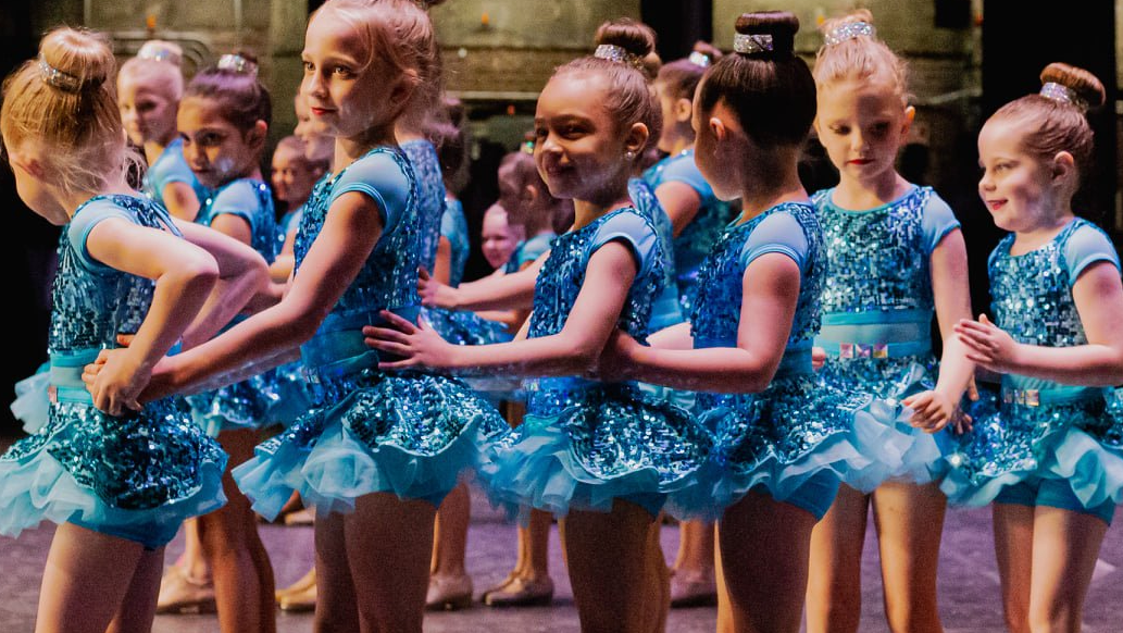 A group of young girls in blue dresses are dancing on a stage.