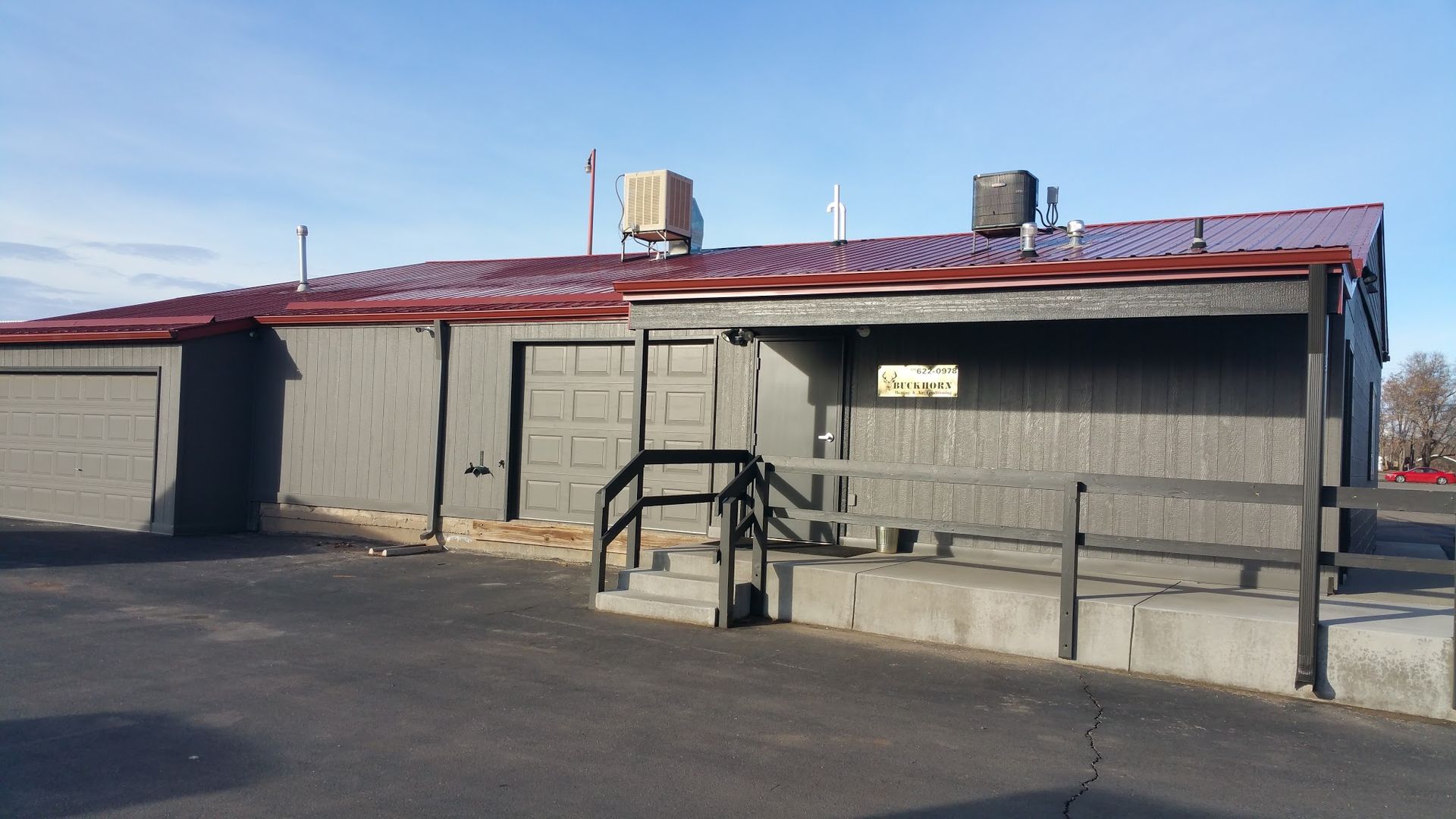 A single-story gray building with a red metal roof, a loading dock, and a small porch ramp under a bright blue sky.