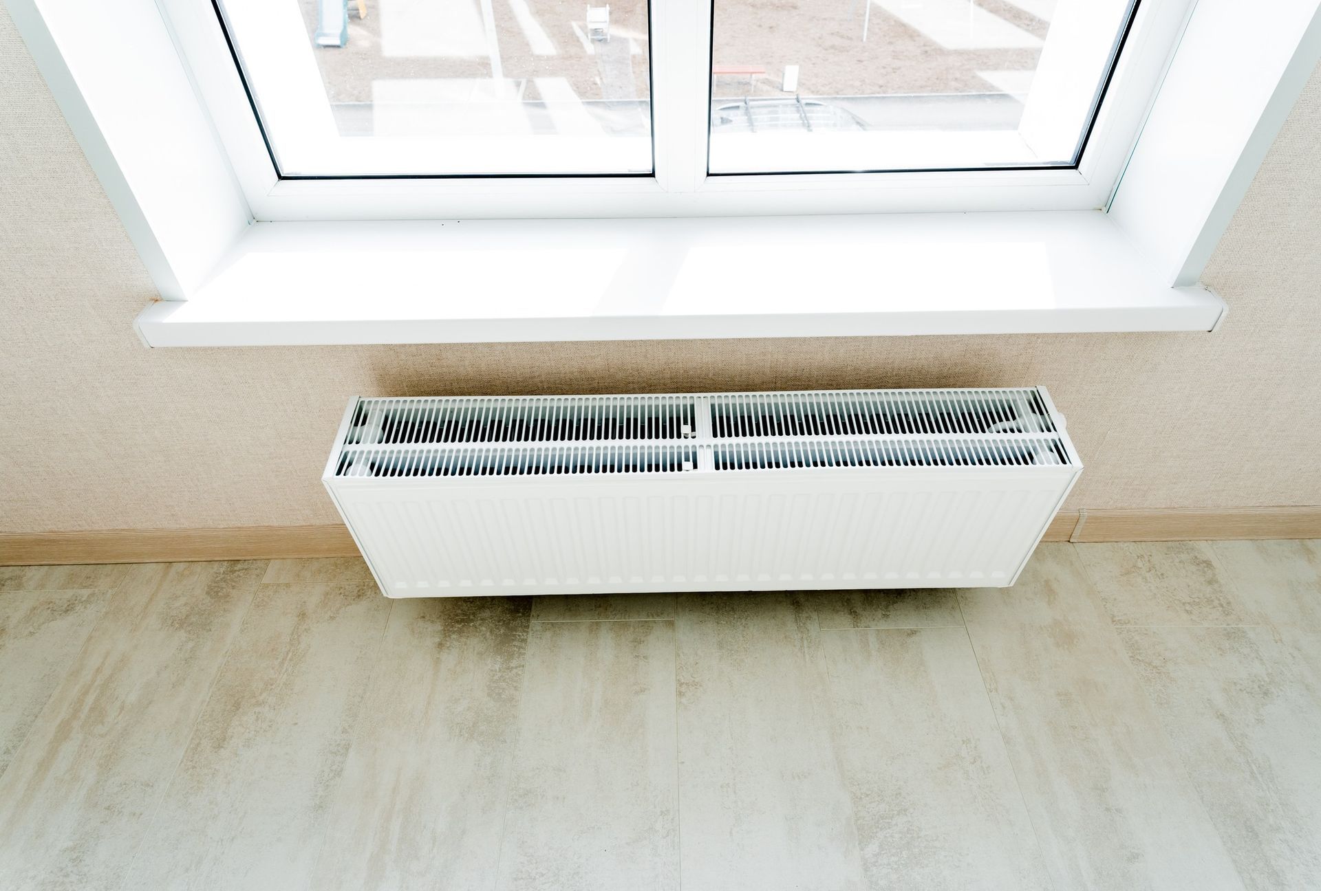 A white radiator mounted on a beige wall beneath a white window sill and a window.